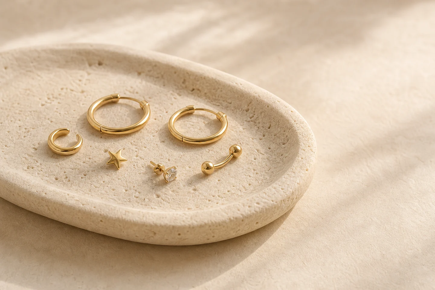 A small collection of gold hoops, a cuff, studs, and a curved barbell on warm limestone